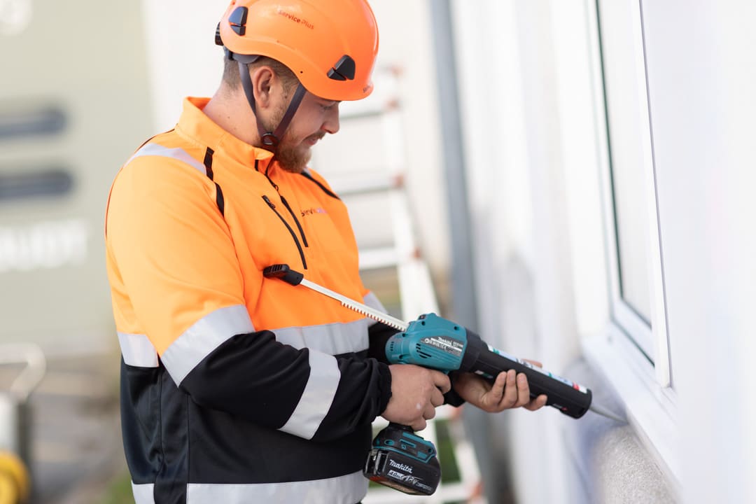 :En håndværker iført sikkerhedshjelm og ServicePlus-arbejdstøj arbejder med fugning omkring et vindue.